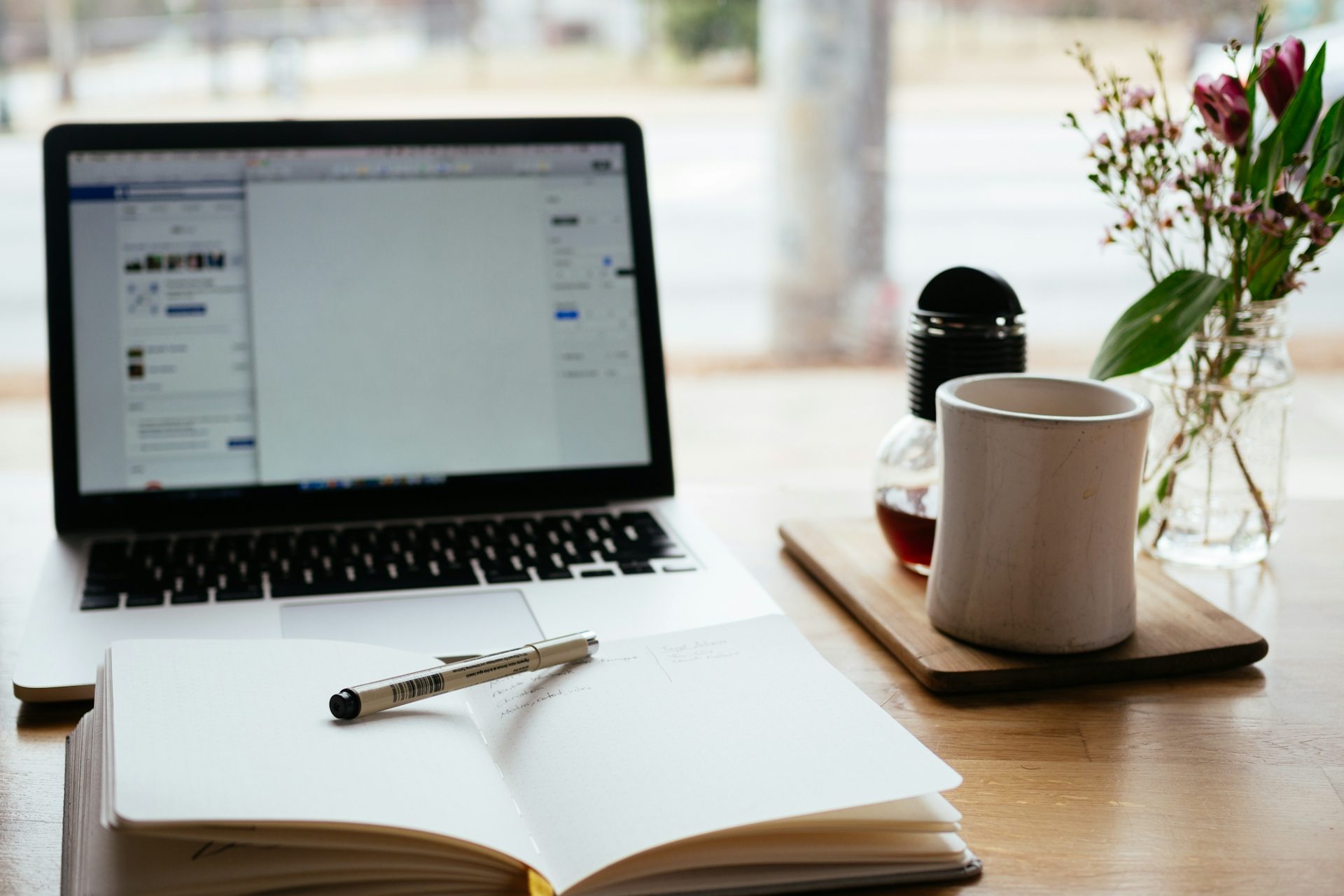 Schreibtisch mit Laptop, Notizbuch, Stift, Tasse Kaffee und frischen Blumen.
